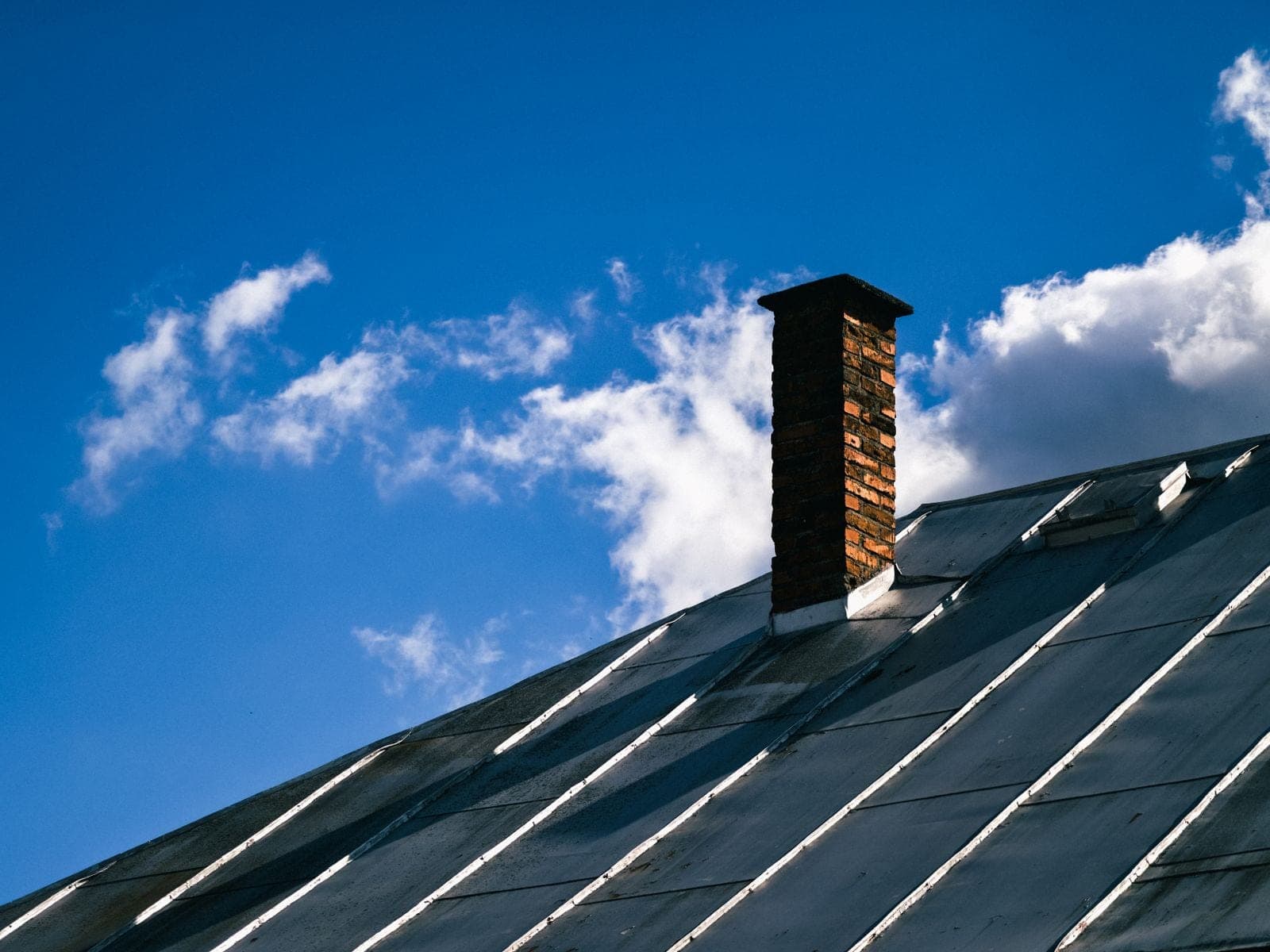 Dark standing seam metal roof with a brick chimney against a blue sky